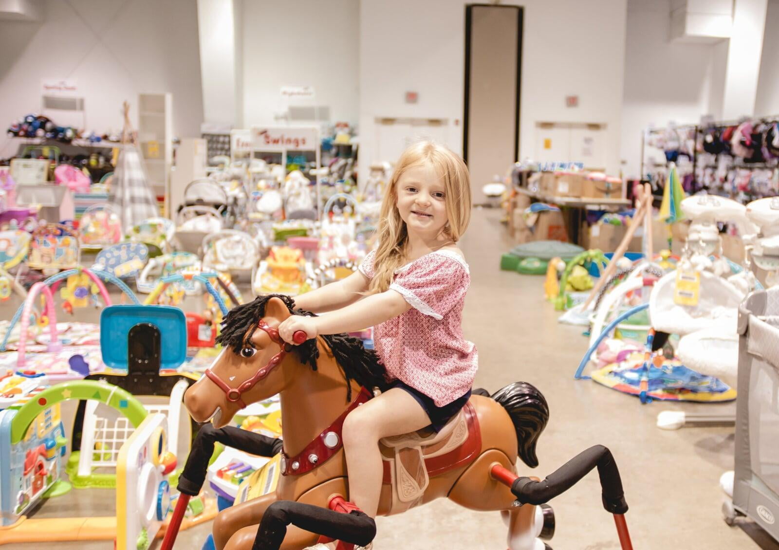 Young girl sits on a rocking horse toy in the toy section at her local JBF sale.